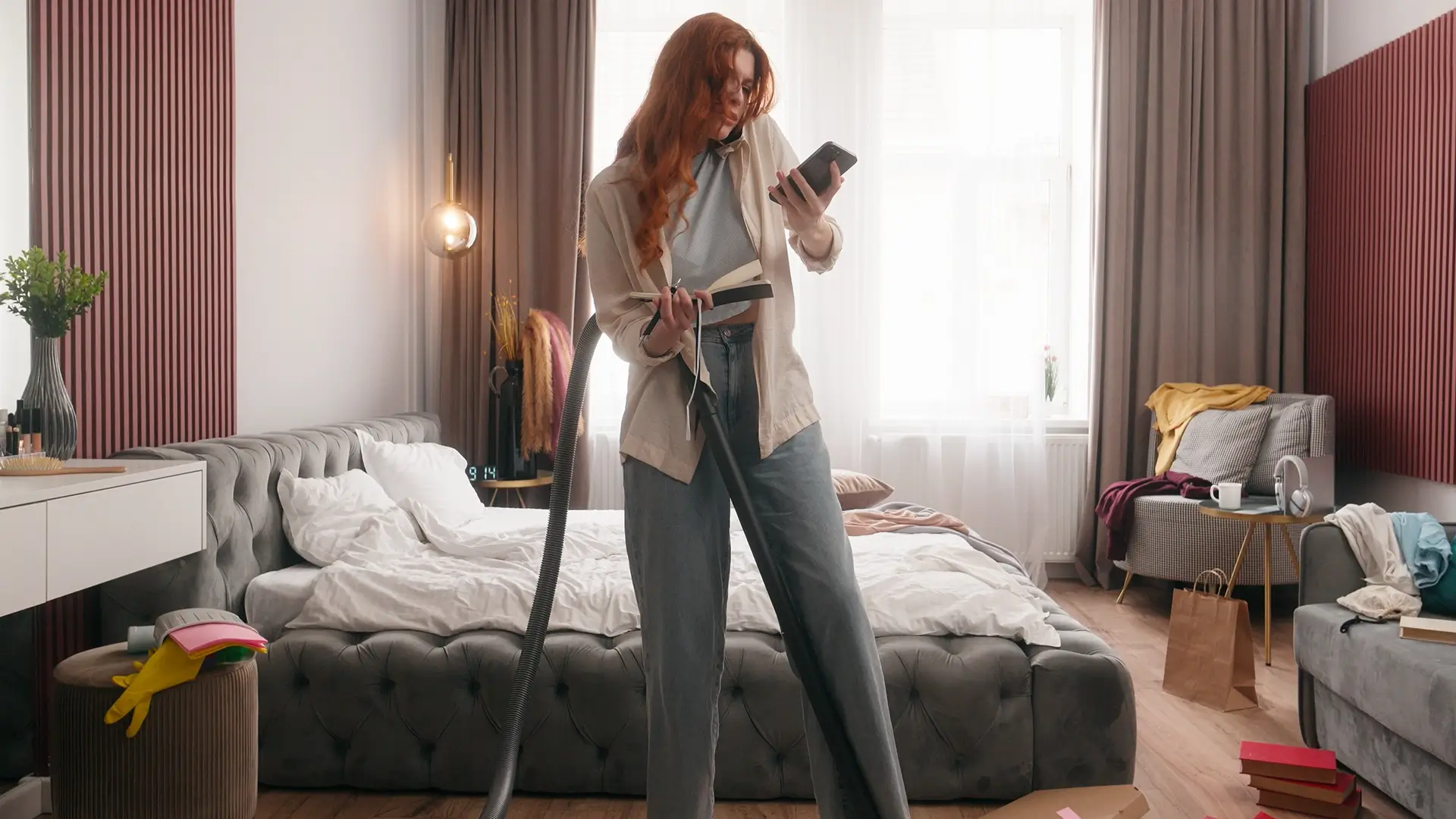 Woman multitasking with phone, book, vacuum in bedroom.